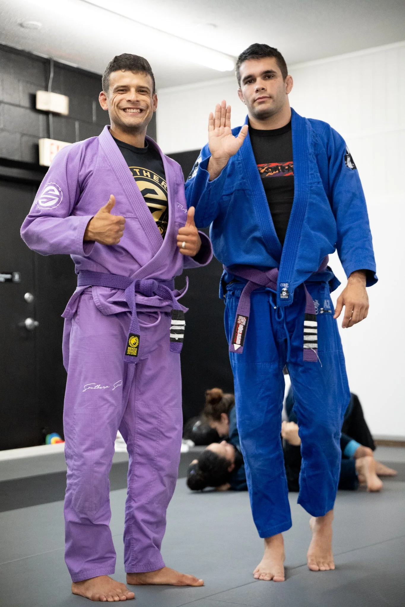 Two men in Brazilian Jiu-Jitsu gis standing on a mat, one in purple and the other in blue, with other practitioners sitting in the background.