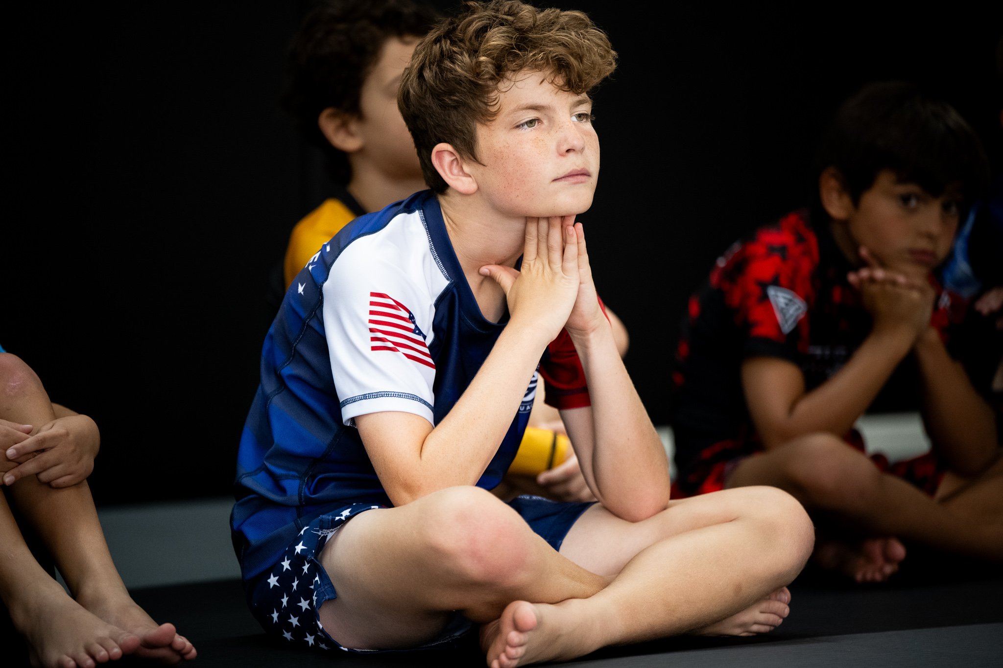 Young boy sitting cross-legged with hands under chin, wearing a sports jersey with American flags, watching attentively against a black background.