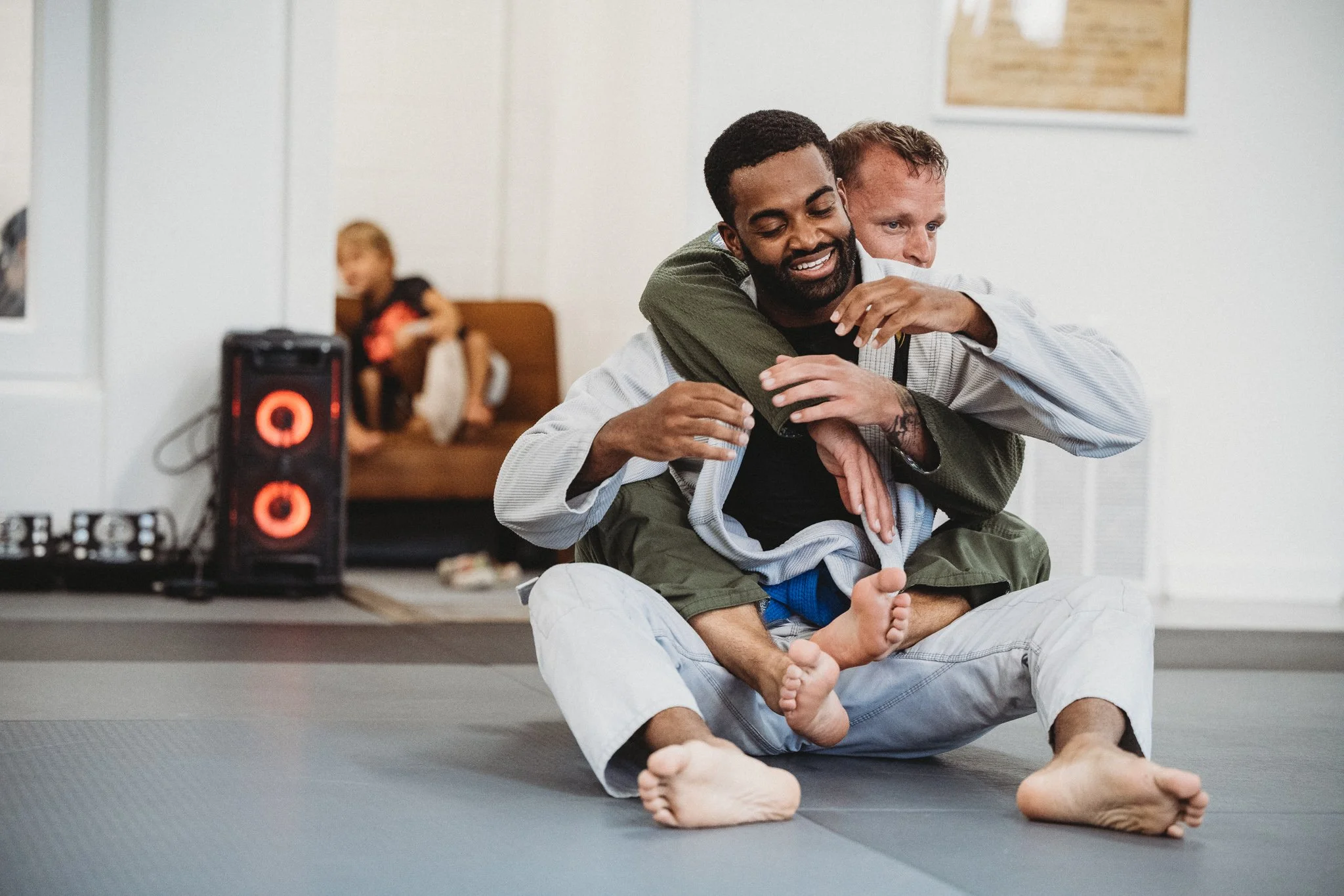 Two men practicing Brazilian Jiu-Jitsu on a gray mat, with a woman sitting in the background.