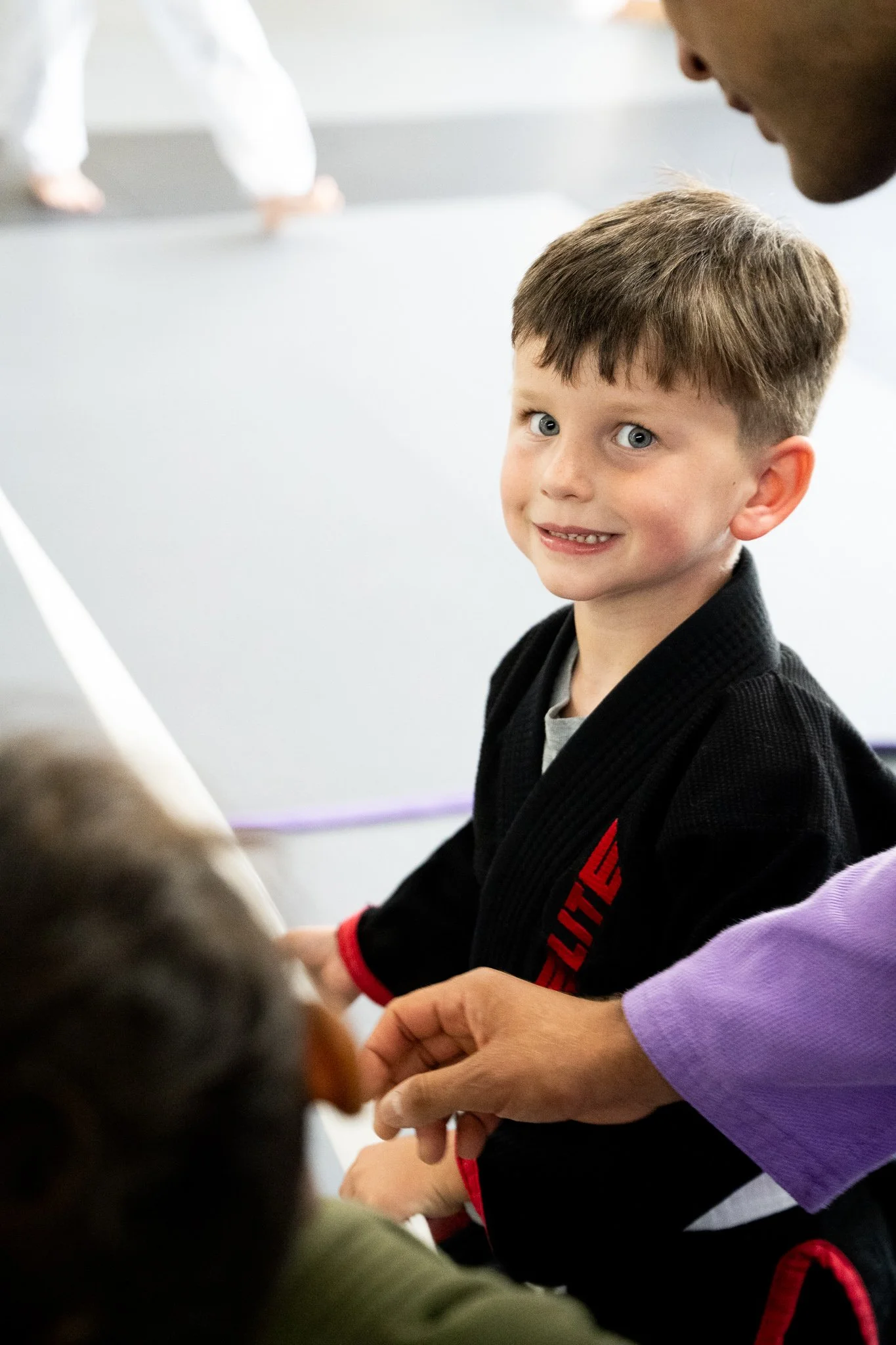 A young boy with short brown hair and blue eyes, wearing a black martial arts gi with red accents, smiling and looking at the camera while holding hands with children in a group activity.
