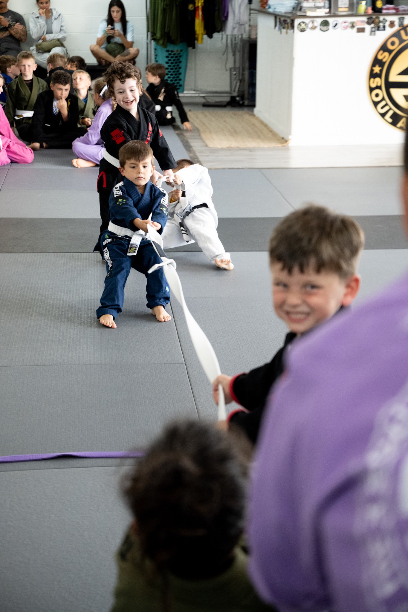 Kids in martial arts uniforms sitting and kneeling on mats, with one child smiling and holding a white belt, in a martial arts dojo.