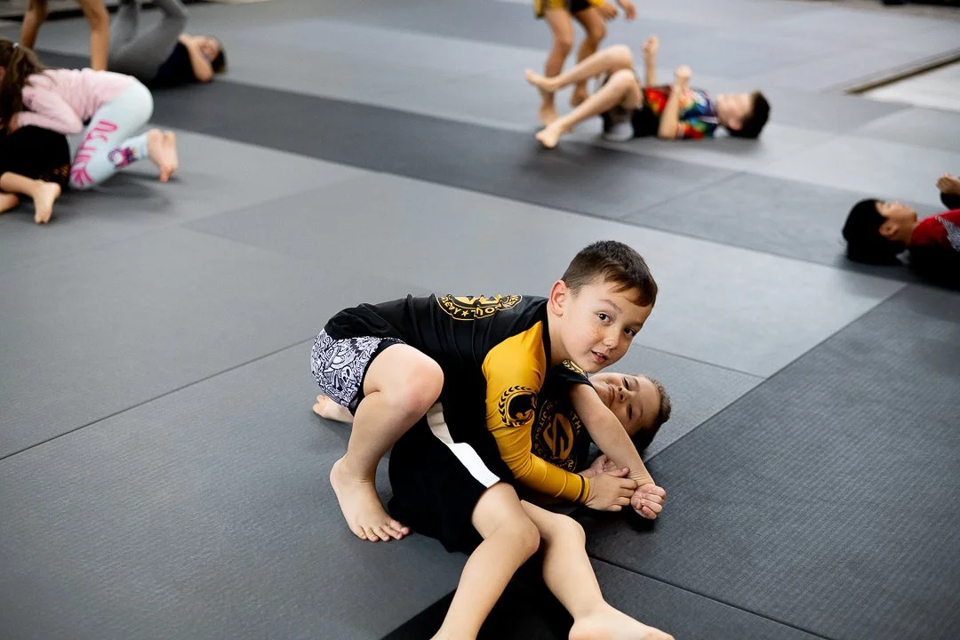 Children practicing Brazilian Jiu-Jitsu on black mats, some are rolling and grappling.