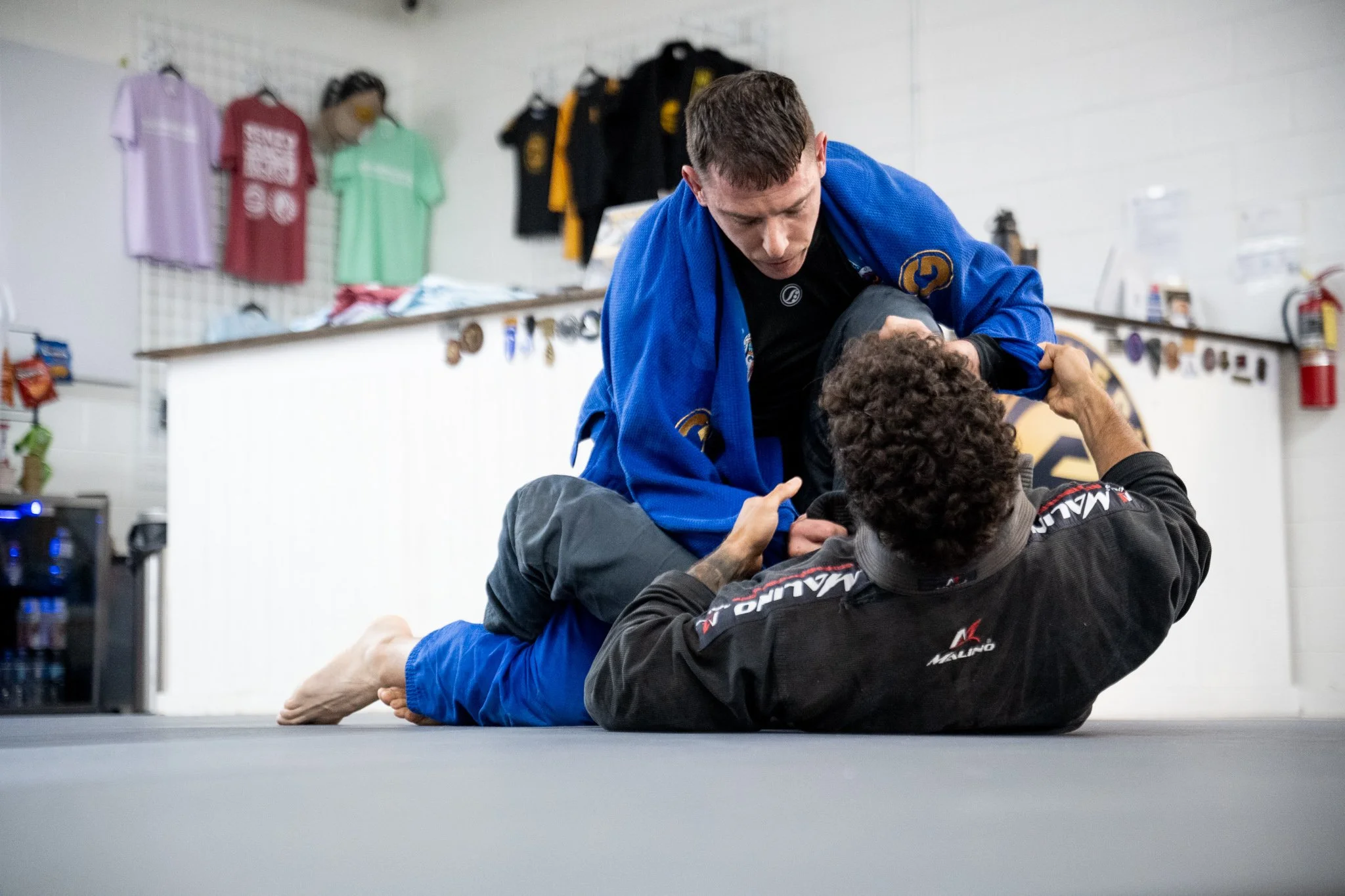 Two people practicing Brazilian Jiu-Jitsu on a gray mat inside a martial arts gym. One person in a blue gi is on top, gripping the sleeve of the opponent's black gi, while the other lays on their back with curly hair. The background shows a wall with hanging colorful martial arts uniforms and equipment.