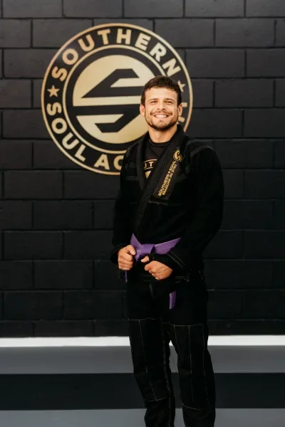 A smiling man in a black Brazilian Jiu-Jitsu gi with a purple belt standing in front of a black wall with a circular logo that reads 'Southern Soul Academy'.