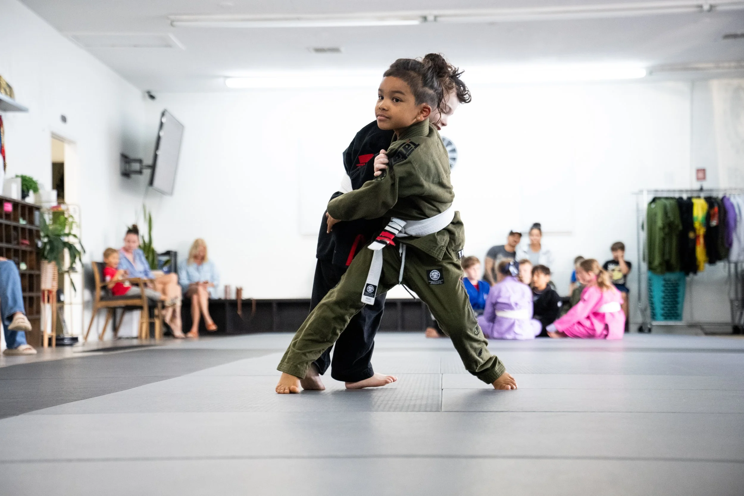 Two children practicing Brazilian Jiu-Jitsu on a gray mat at a martial arts class, with onlookers seated in the background.