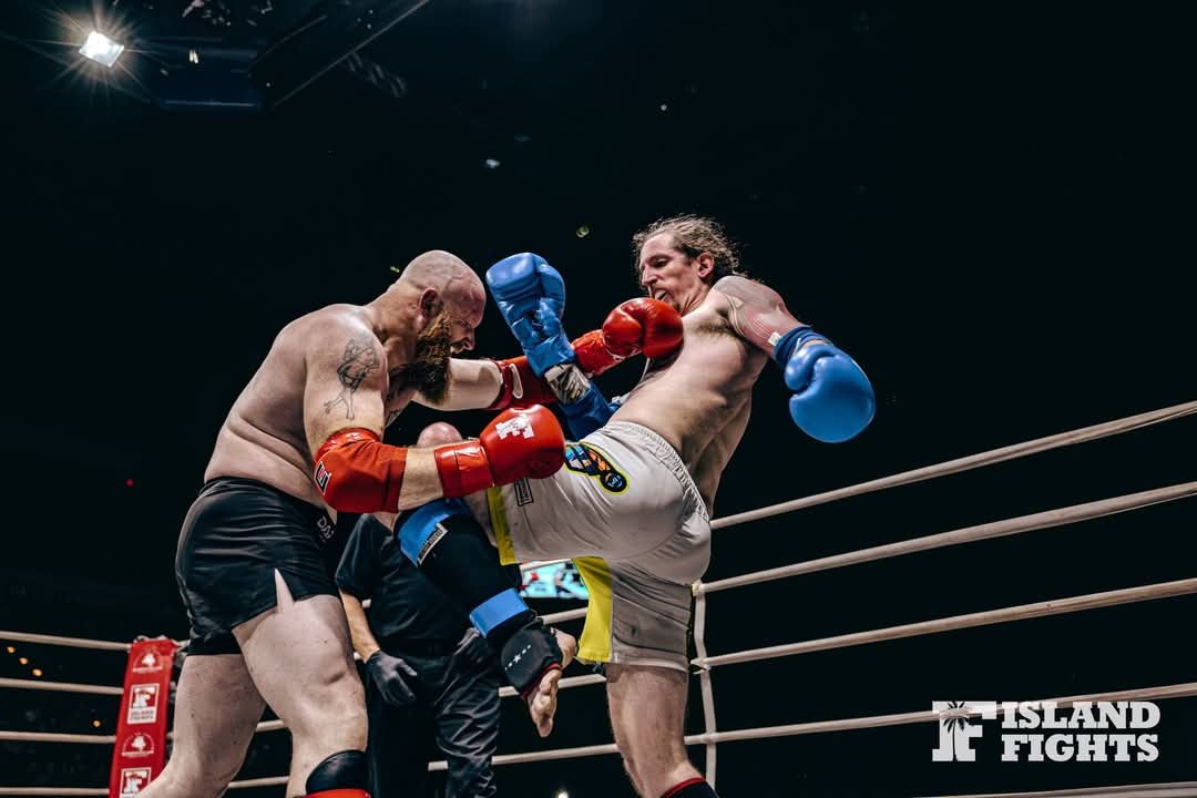 Two male fighters wearing boxing gloves, one in red and the other in blue, engaged in a kickboxing match inside a ring with a black background. One fighter is delivering a punch to the other's face, while the second fighter is mid-kick. An referee observes the fight.