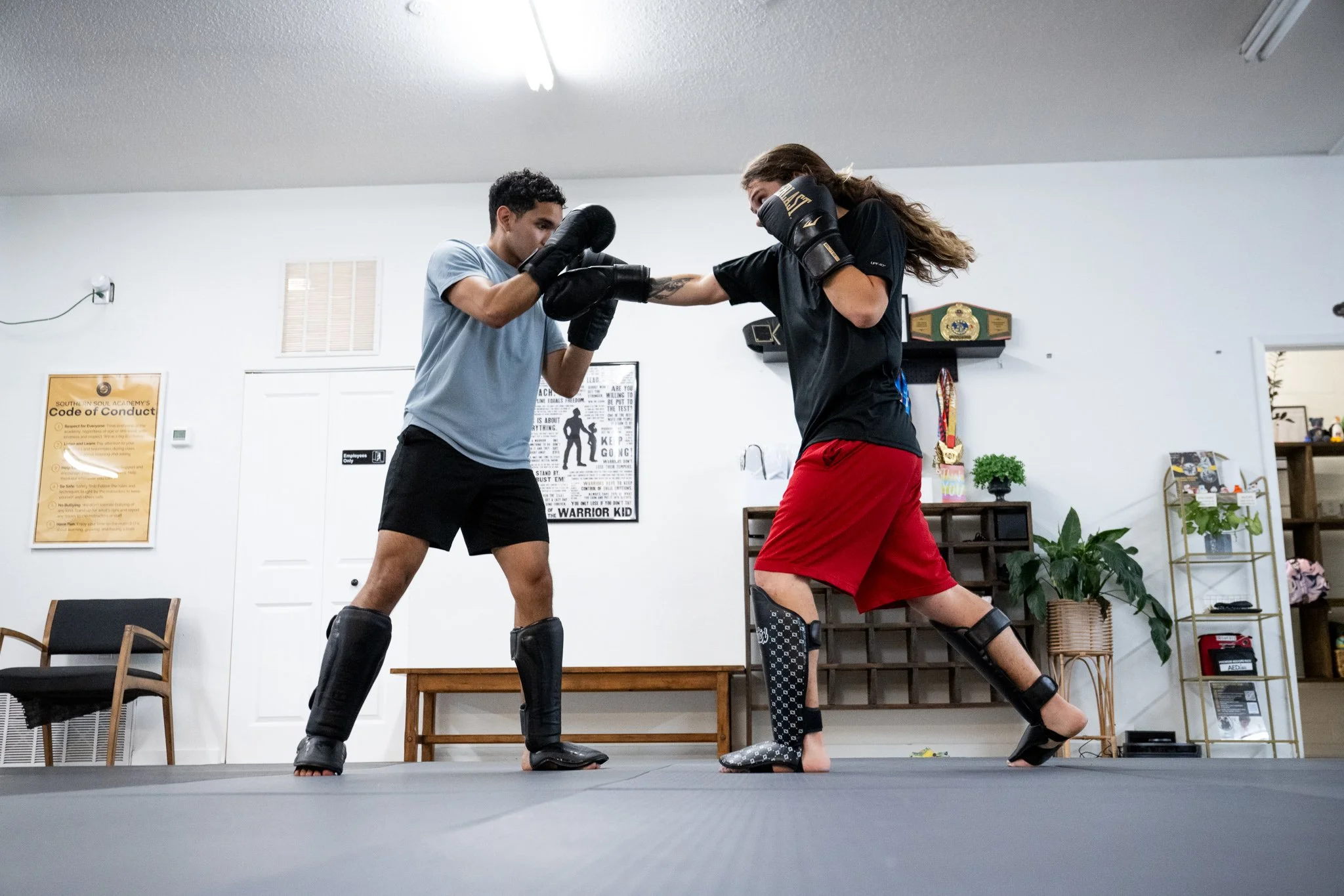 Two people practicing boxing in a gym, wearing boxing gloves and protective gear, engaged in a sparring match on a padded mat.
