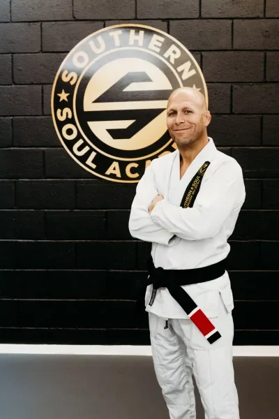 A man in a white Brazilian Jiu-Jitsu gi with a black belt standing in front of a black wall and a logo that reads 'Southern Soul Academy'. he is smiling with arms crossed.