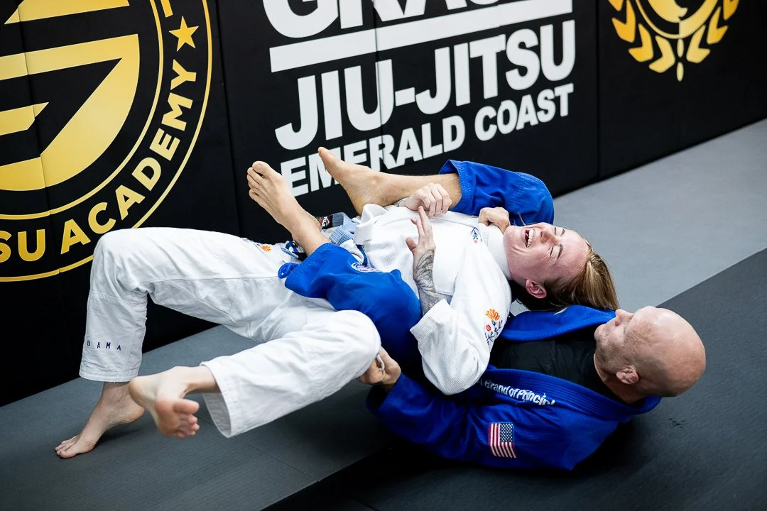 Two women practicing Brazilian Jiu-Jitsu on mats, one in a white gi and the other in a blue gi, with a black wall featuring yellow and white branding in the background.