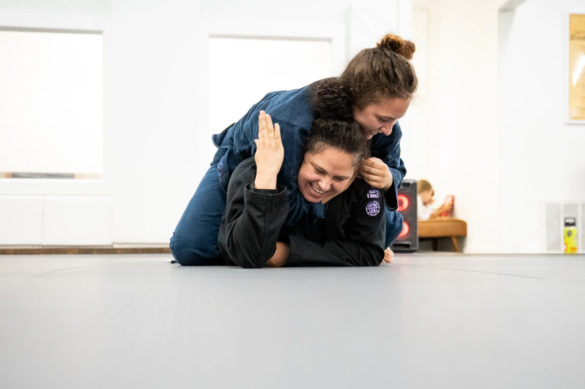 Two women practicing Brazilian Jiu-Jitsu on the floor in a martial arts gym, smiling and engaged in a friendly sparring match.