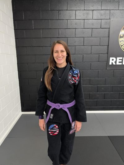 Young woman in a black Brazilian Jiu-Jitsu gi with purple belt, standing on a gray mat in a martial arts training facility with black and white walls, smiling at the camera.