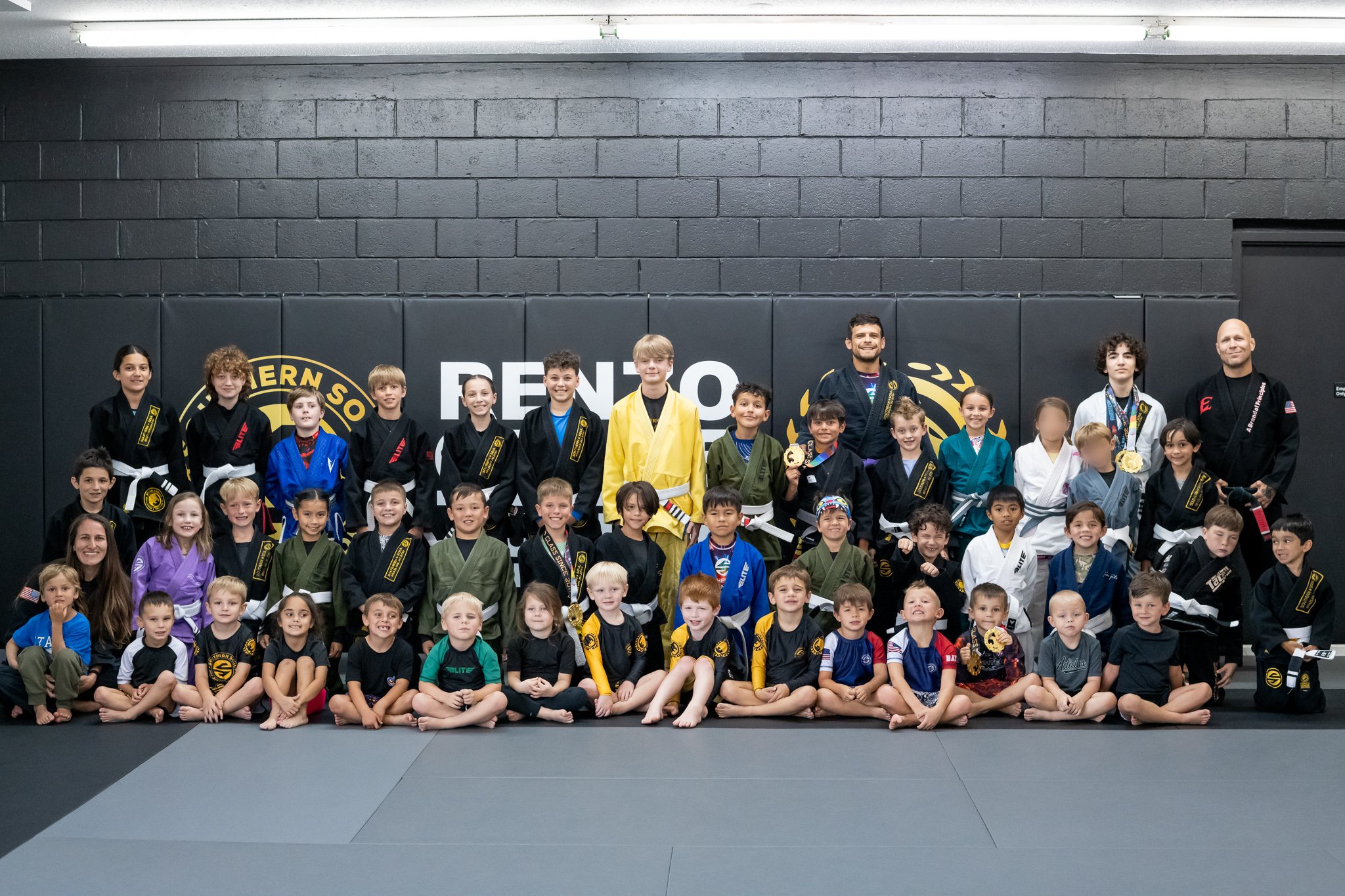 Group of children and adults in martial arts uniforms, some with medals, posing on a competition mat in front of a black wall with the words 'RENO' and a logo, inside a martial arts training facility.