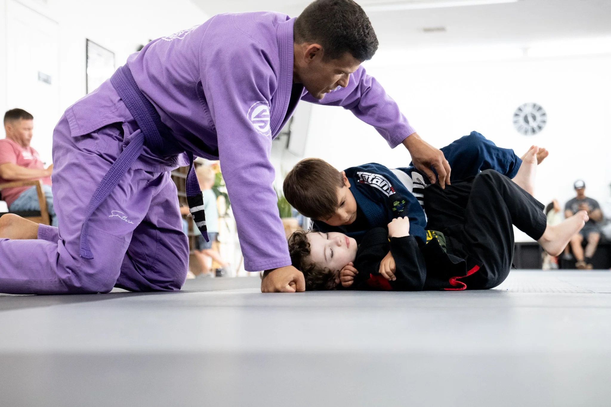 A Brazilian Jiu-Jitsu instructor demonstrates a grappling technique on a young student during a class. The instructor wears a purple gi and the student wears a black gi. Other students and observers are visible in the background, seated and watching.