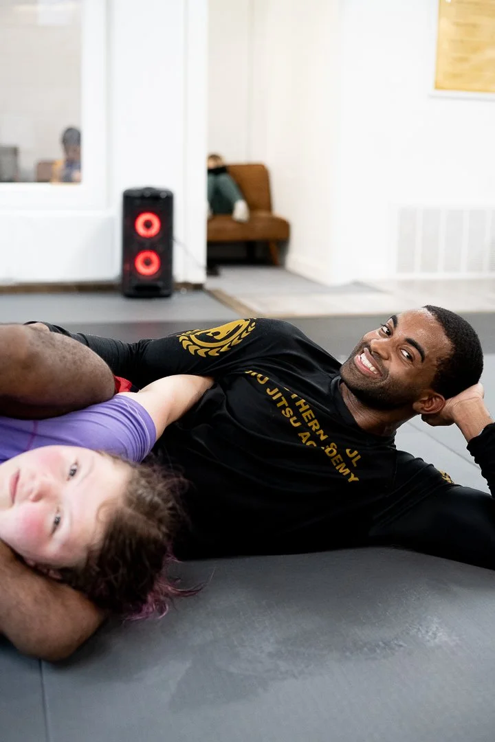 Two people practicing martial arts or Brazilian Jiu-Jitsu on a mat, with one person smiling at the camera.