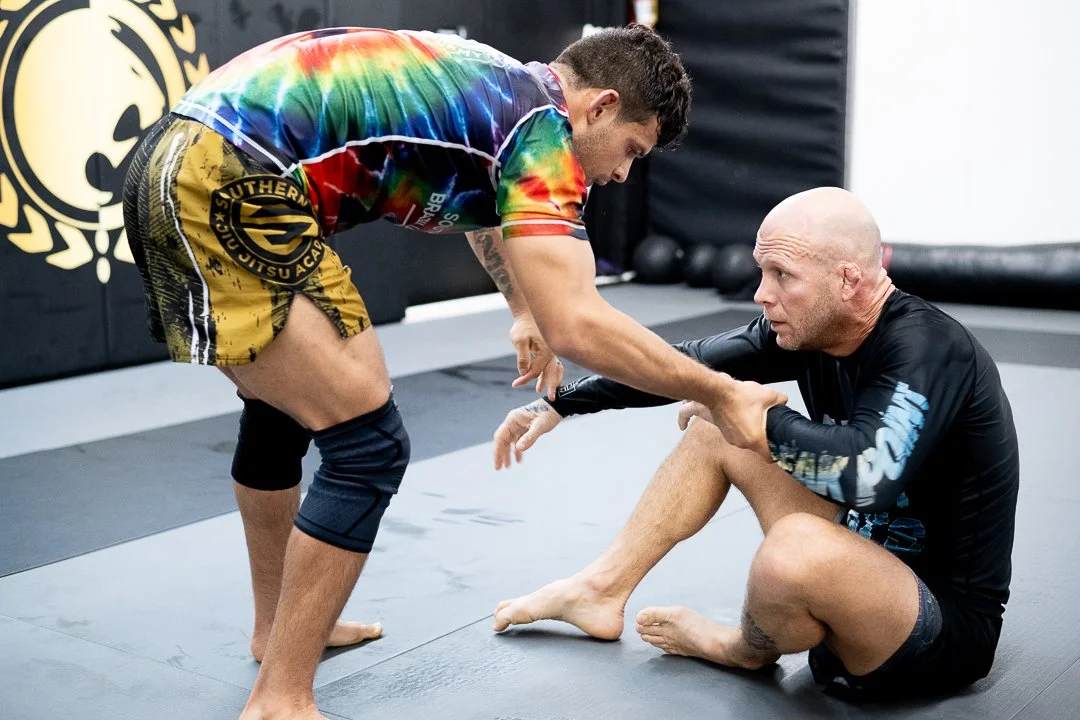 Two men practicing Brazilian Jiu-Jitsu in a gym, one standing and holding the other's arm, who is sitting on the mat.