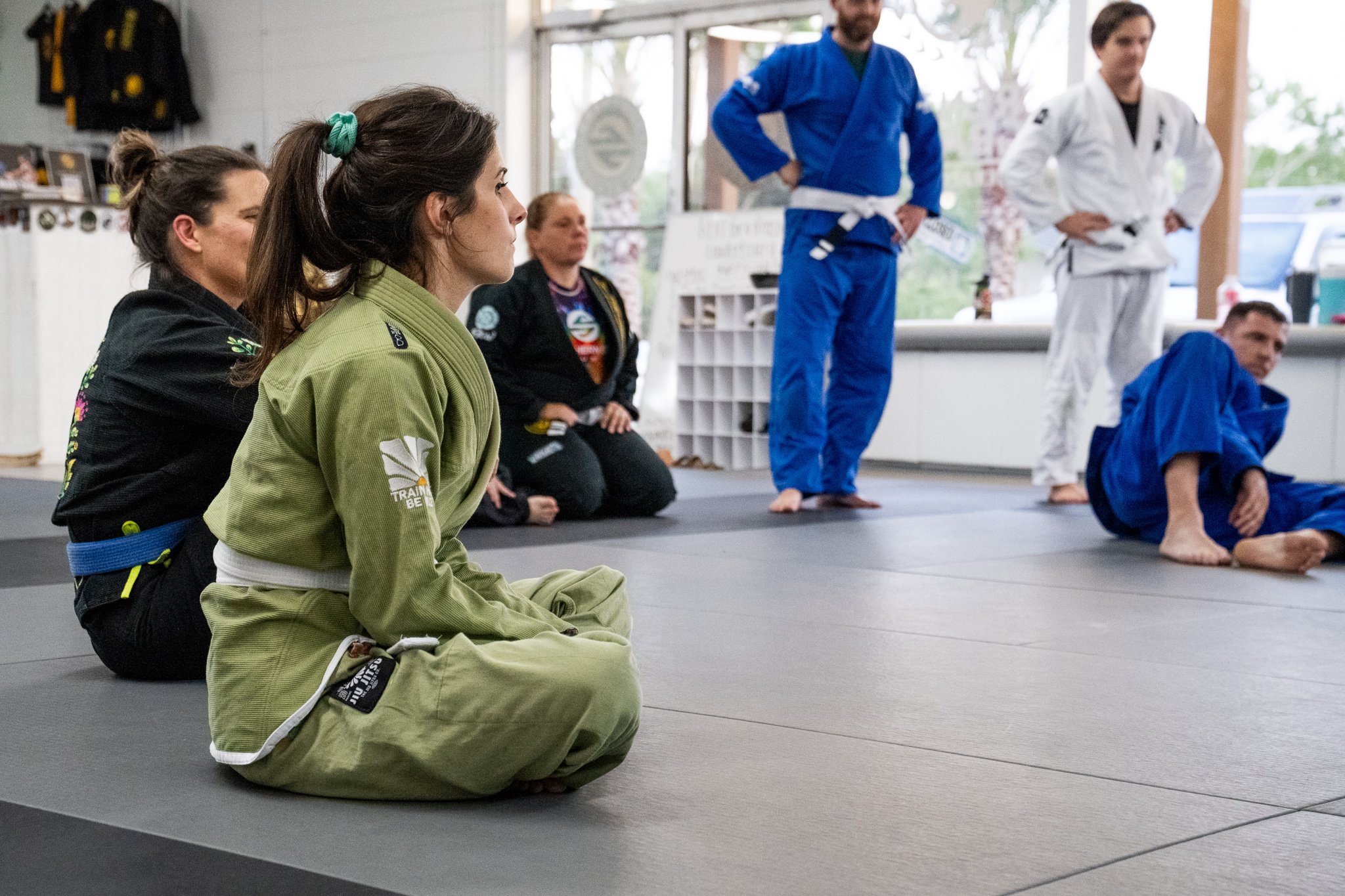 Martial arts class with students wearing gis, sitting on mats, and two instructors standing in the background. One instructor is in a white gi, and the other in a blue gi.