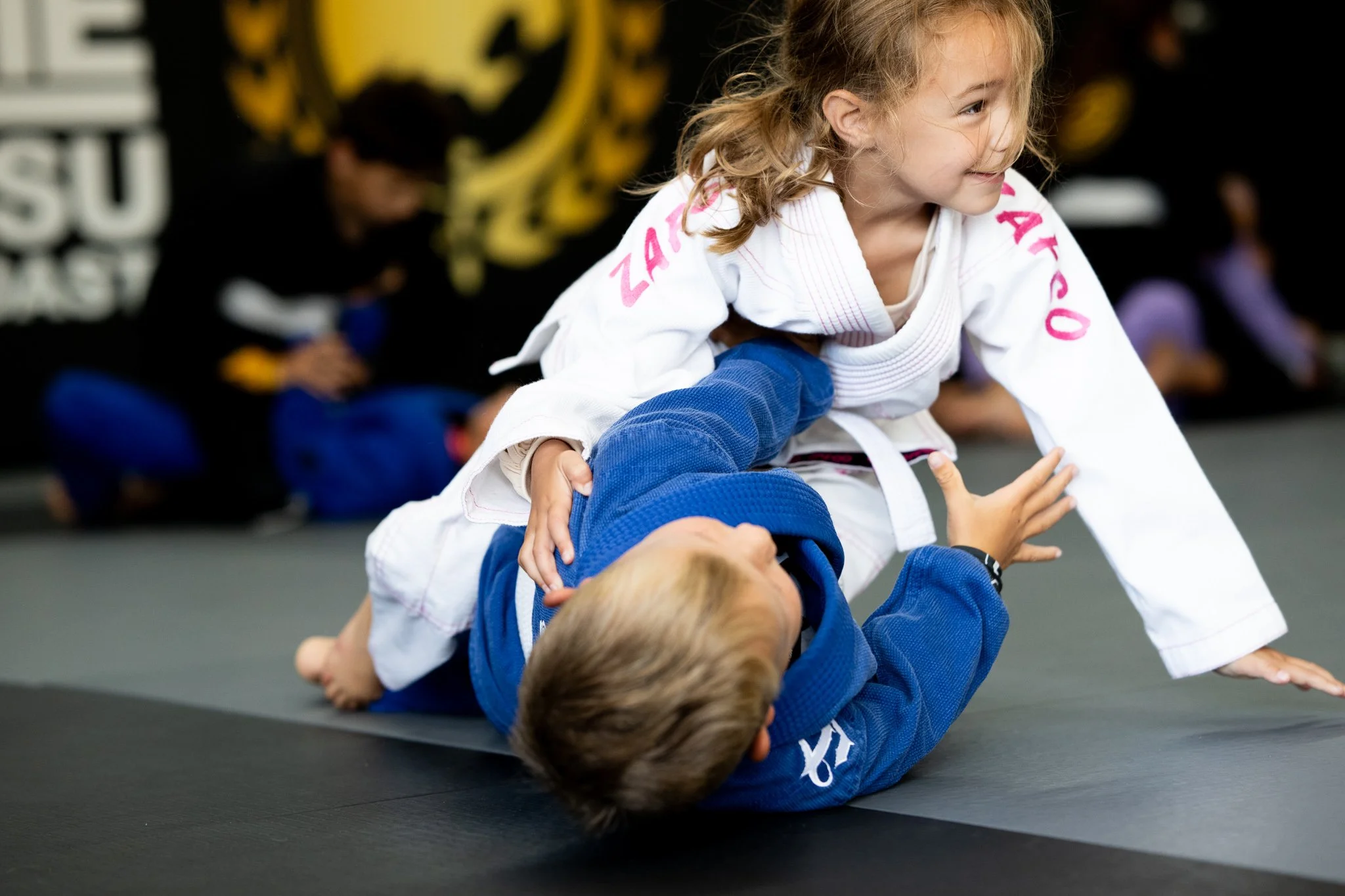 Children practicing Brazilian Jiu-Jitsu on a black mat in a martial arts gym; a young girl in a white gi with pink lettering is executing a move on a boy in a blue gi who is on his back.