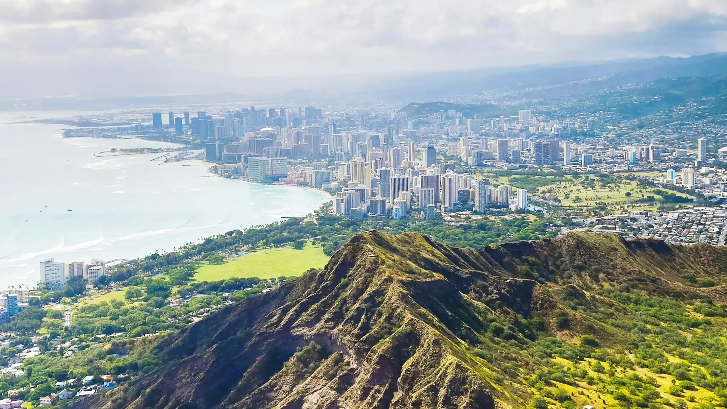 Aerial view of a cityscape with high-rise buildings, a coastline, and lush green parks seen from a mountain.