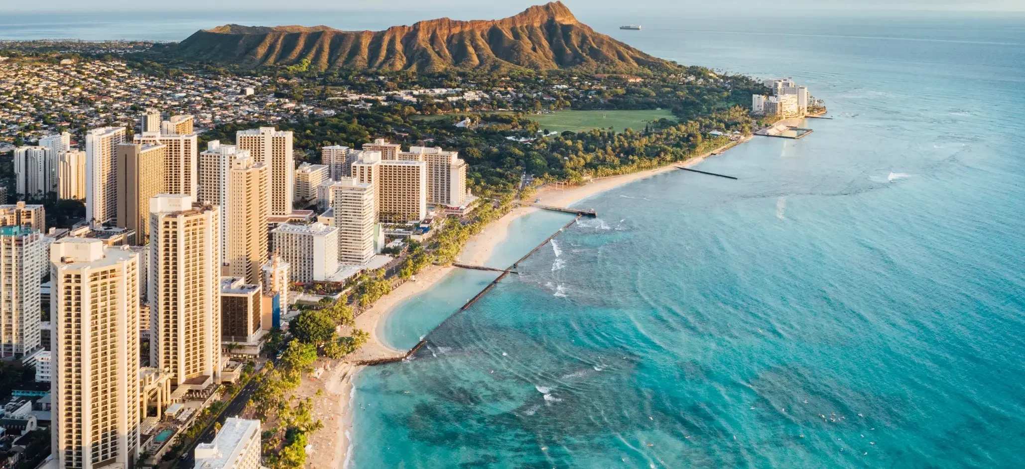 Aerial view of a coastal city with high-rise buildings along the beach, with Diamond Head volcanic crater in the background and the ocean extending to the horizon.