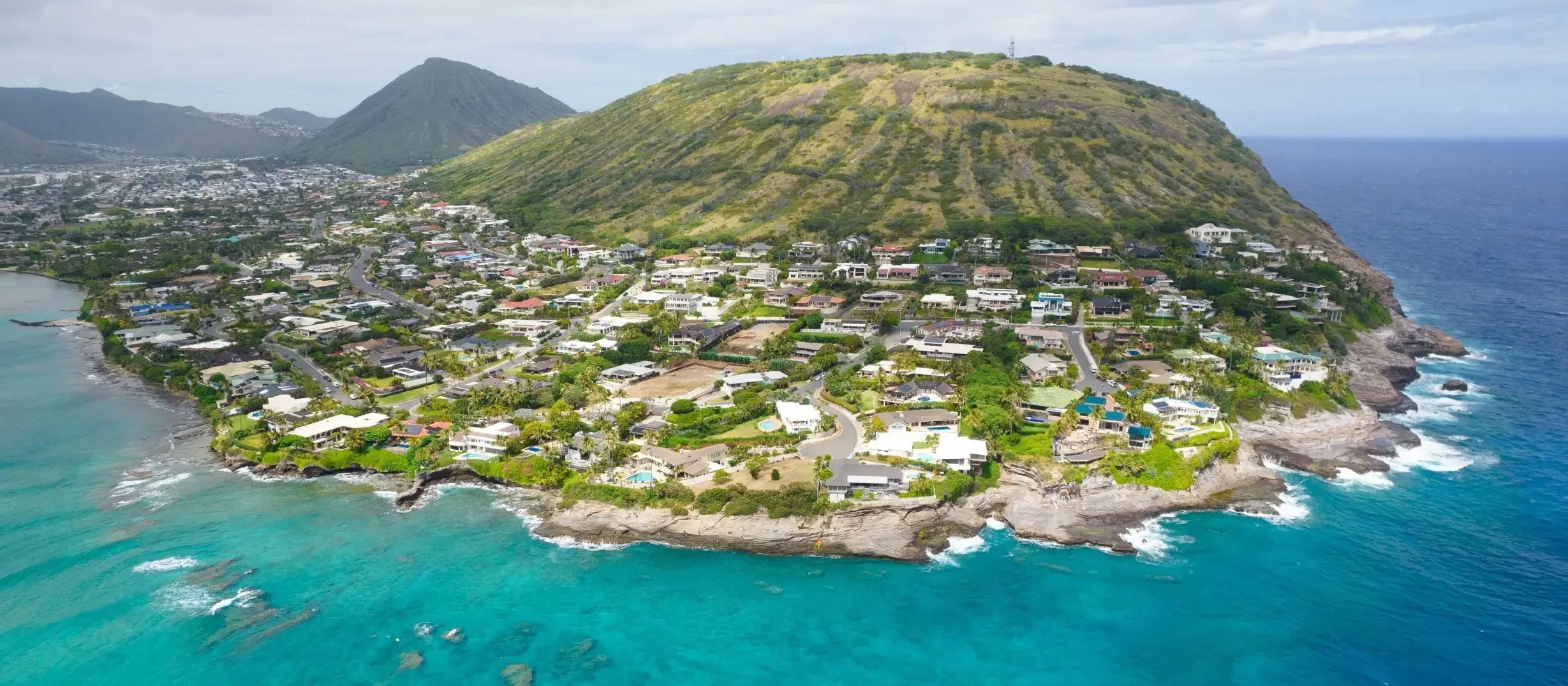 Aerial view of a coastal residential area surrounded by green hills and blue ocean waters.