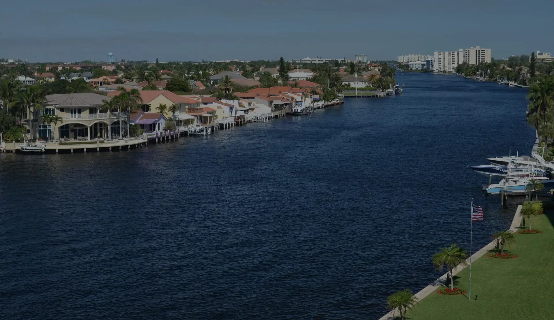 A scenic view of a residential neighborhood along a waterway with boats docked at private docks, tall apartment buildings in the distance, and an American flag in the foreground.
