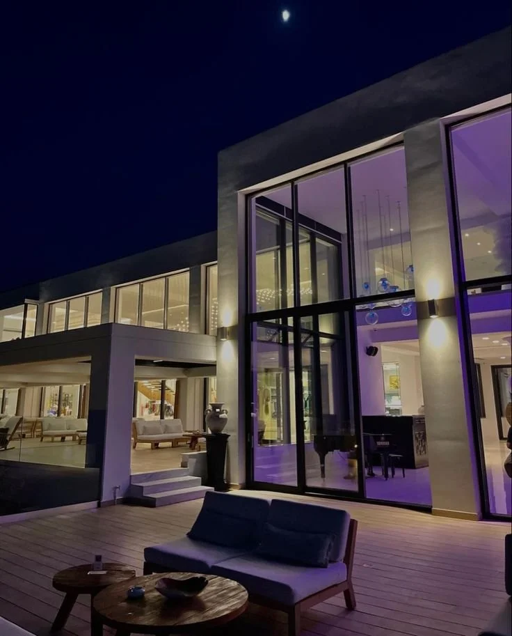 Modern house exterior at night with large glass windows, illuminated interior, outdoor seating area, and a visible moon in the sky.
