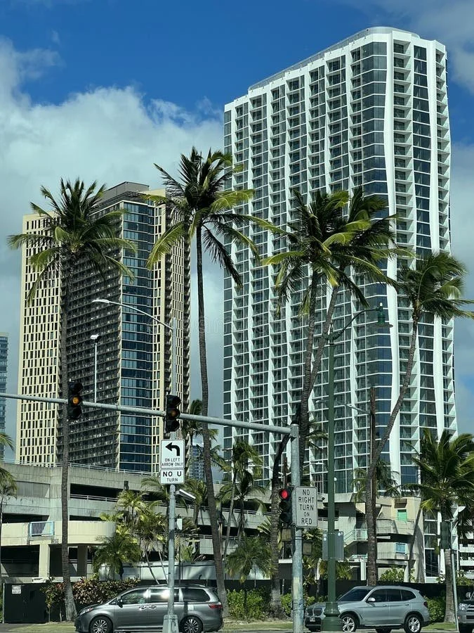 Tall modern high-rise buildings with glass windows, surrounded by palm trees, under a partly cloudy sky, with cars and traffic signals in the foreground.