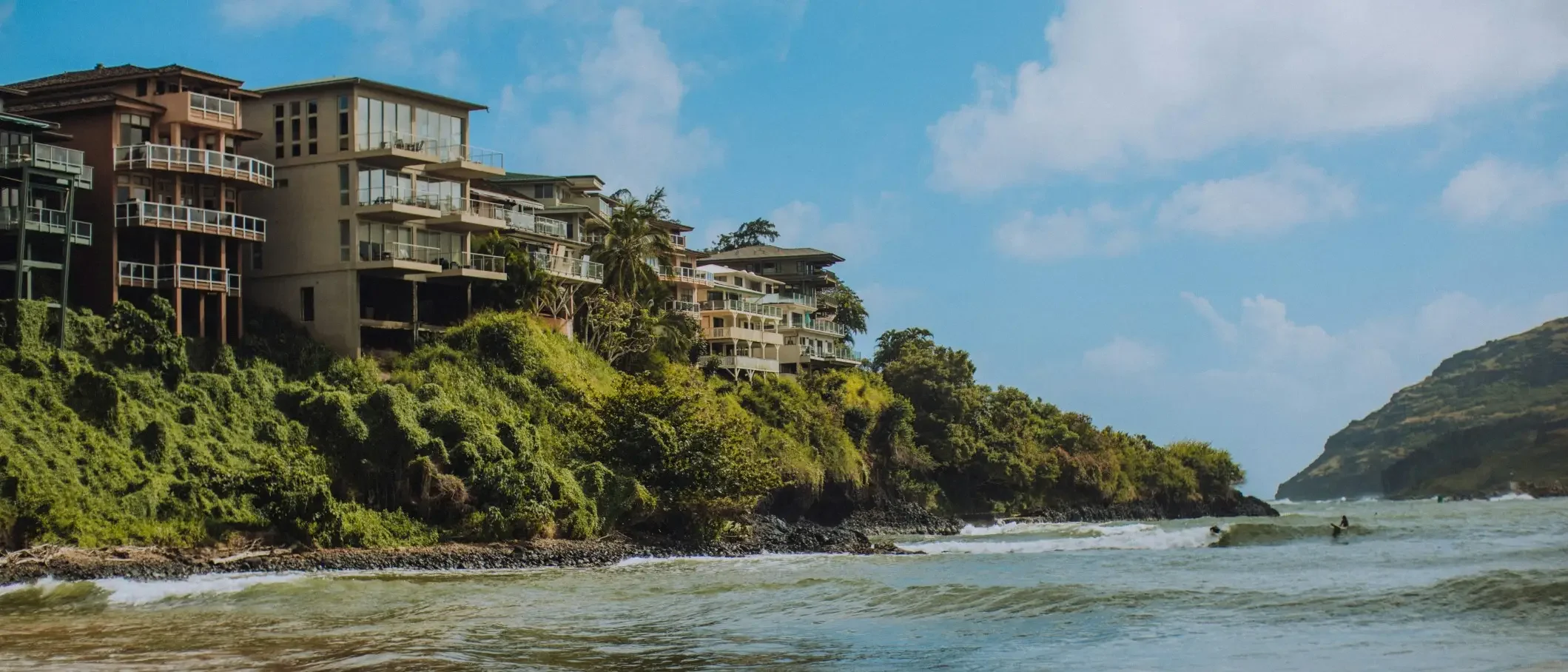 Multiple modern houses built on a lush green hillside overlooking the ocean with blue sky and scattered clouds in the background.