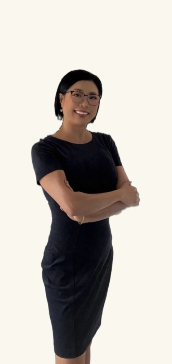 A woman with black hair, glasses, and earrings, smiling with arms crossed, wearing a black dress, standing against a plain light background.