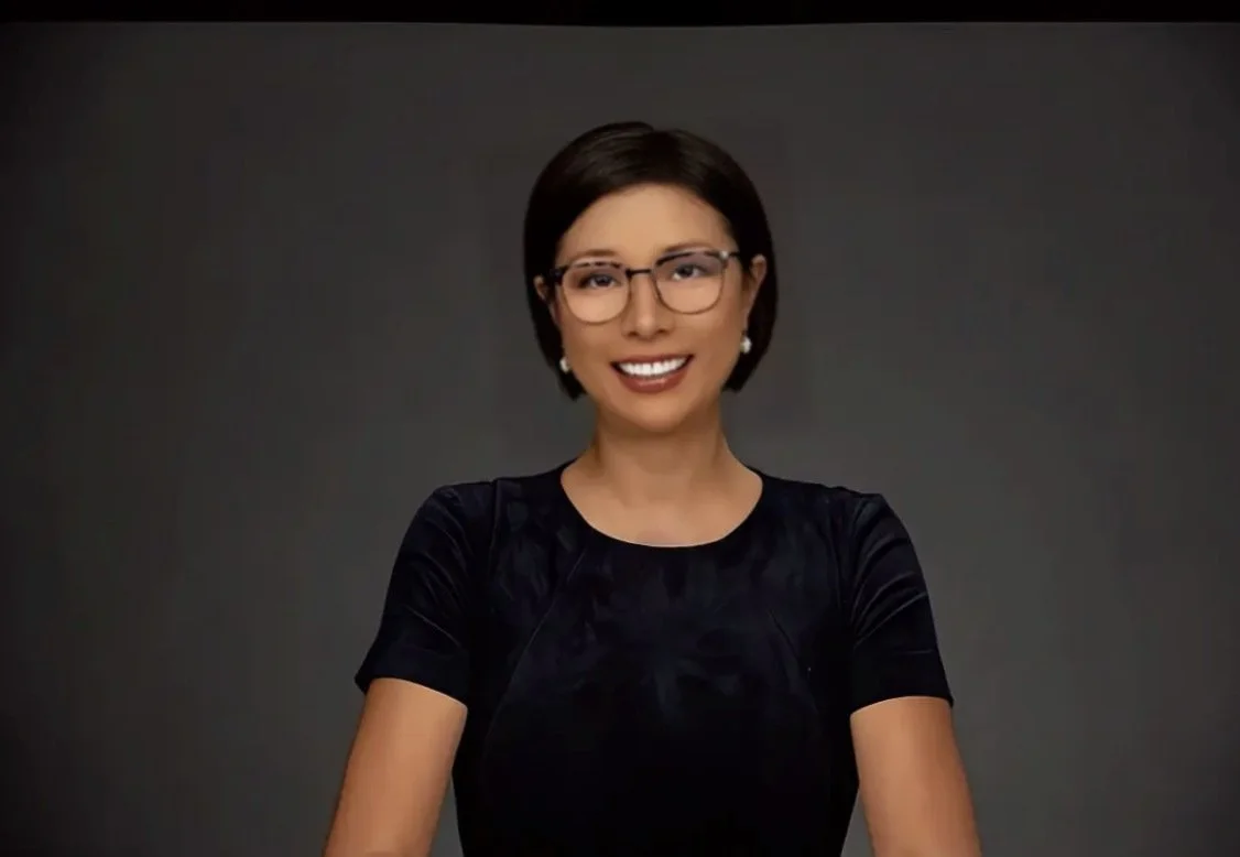 A woman with short dark hair, wearing glasses and a black top, smiling against a dark background.
