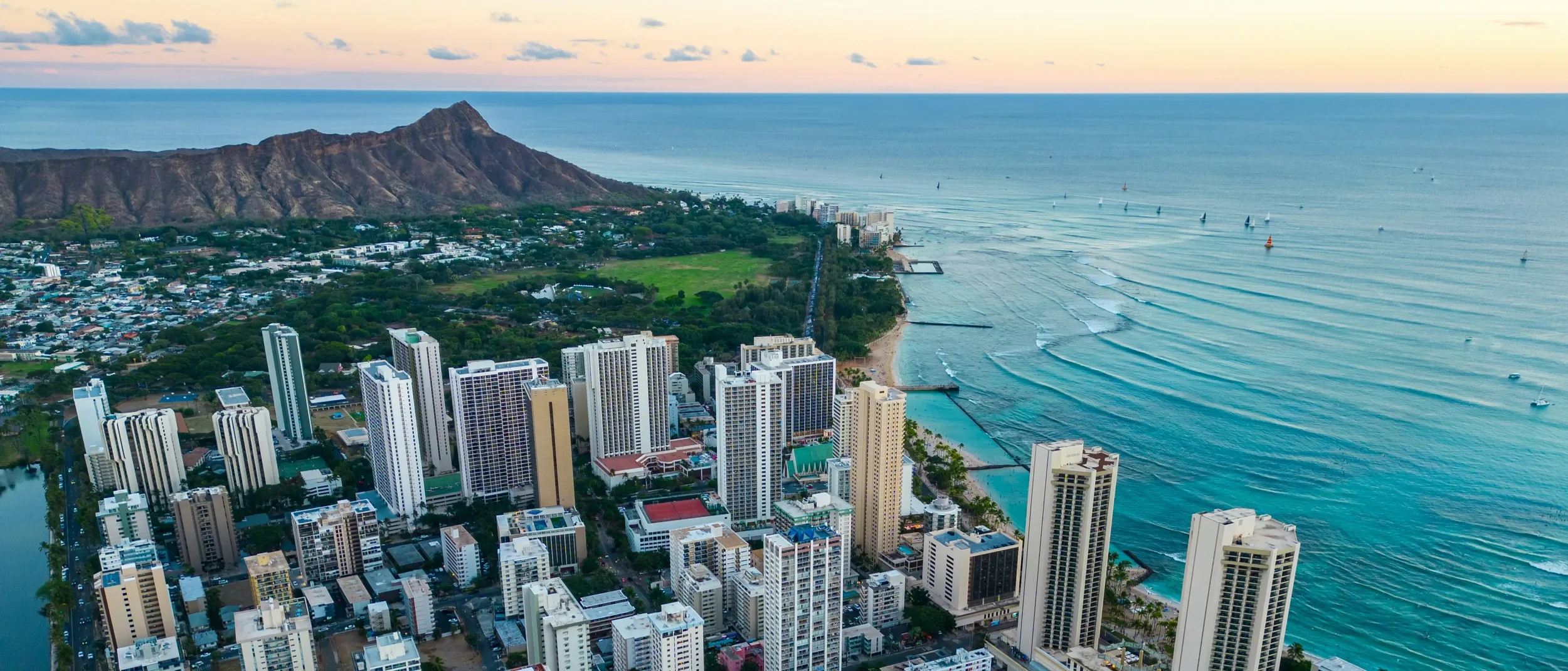 An aerial view of Waikiki beach with high-rise hotels, a green park, and Diamond Head mountain in the background during sunset.