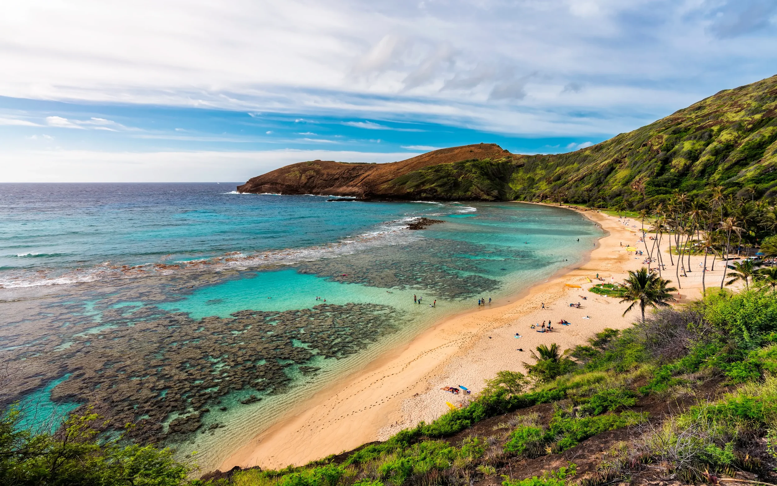 A tropical beach with clear turquoise water, white sand, palm trees, and a lush green hillside in the background under a blue sky with clouds.
