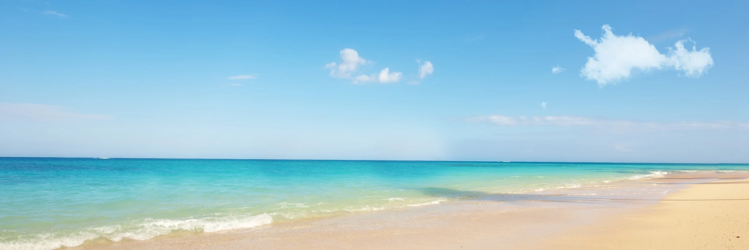 A serene beach with clear turquoise water, a sandy shore, and a bright blue sky with scattered white clouds.
