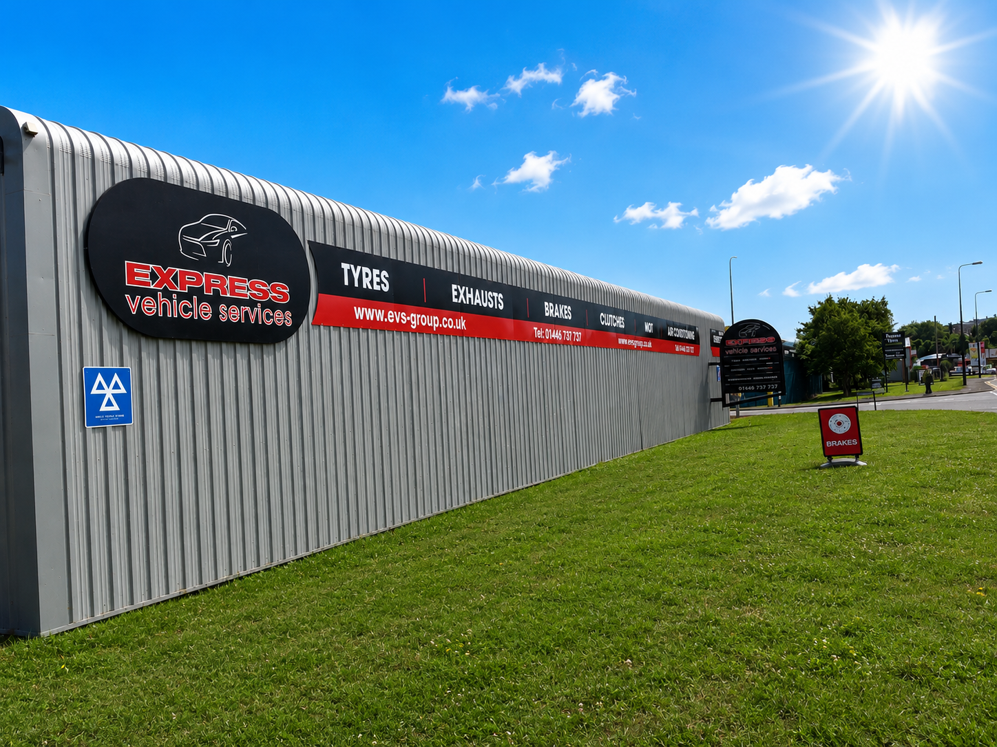 Exterior of an auto repair shop with a large sign displaying services such as tires, exhausts, brakes, clutches, MOT, and oil service. The sign includes the website www.evs-group.co.uk and phone number 01446 737 737. The shop has a metal exterior, a bright sunny sky with some clouds, and a grassy area in the foreground.