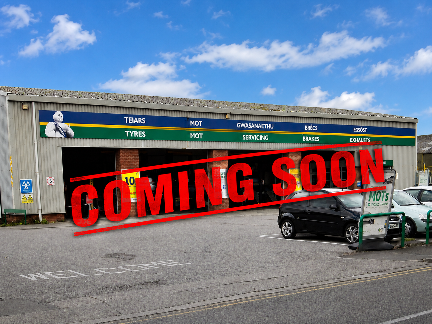 Auto repair shop with a sign displaying services in Welsh and English, a Michelin mascot, and a large red 'COMING SOON' overlay. There are parked cars and a welcoming message painted on the sidewalk.