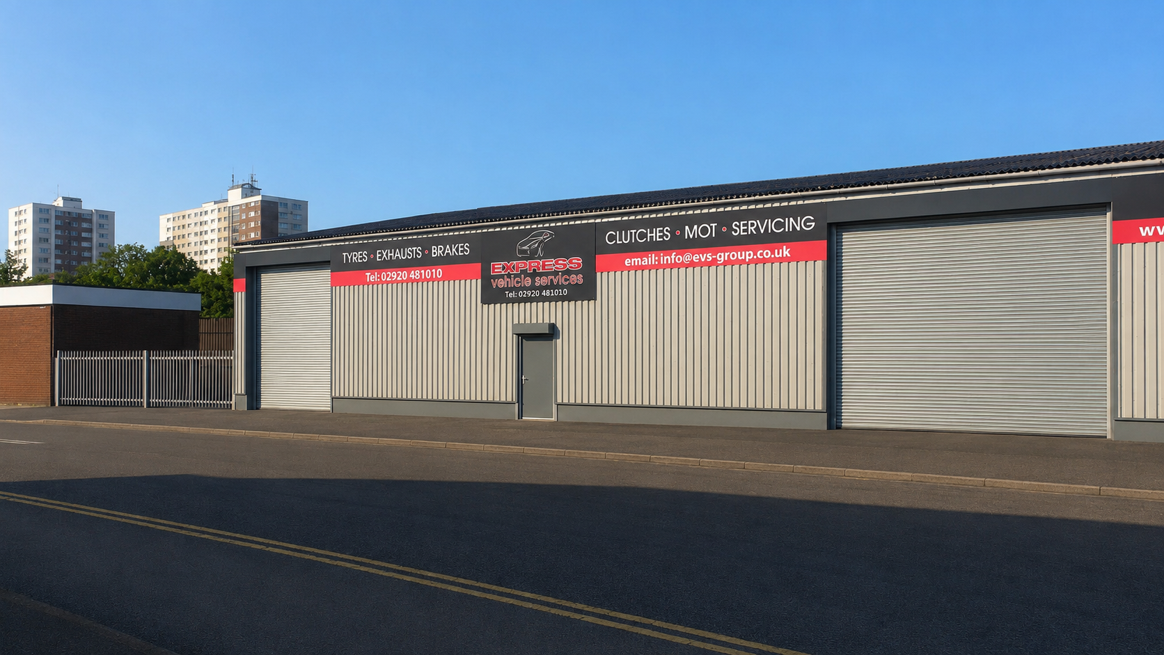 An industrial building with a sign for Express Vehicle Services, offering tyre, exhaust, brake, clutch, and mot servicing. The building has metal roll-up doors and a single pedestrian door, with a street in front and tall apartment buildings in the background.