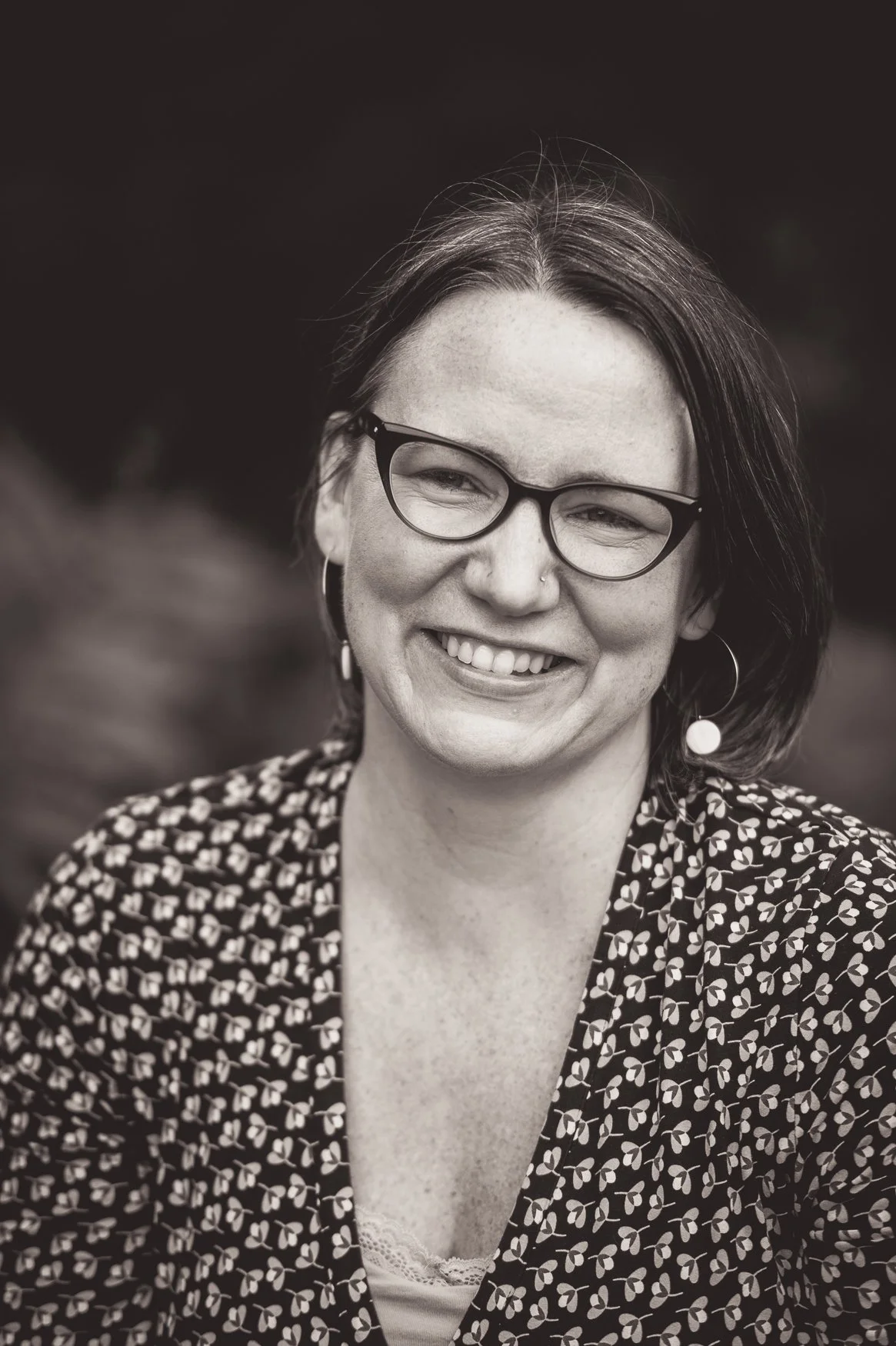 Black and white portrait of a woman with short hair, glasses, and earrings, smiling outdoors.