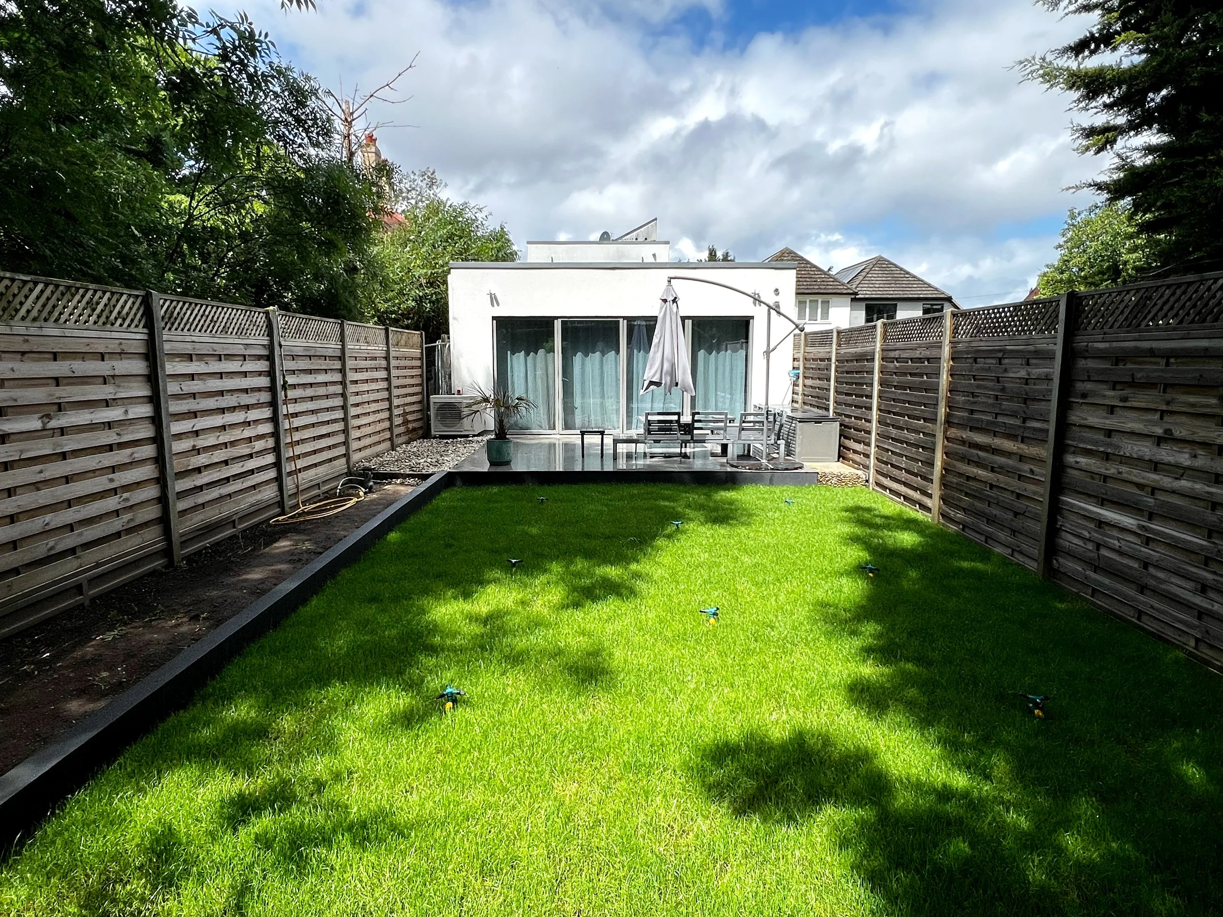 A backyard with a neatly trimmed green lawn, surrounded by wooden privacy fences on both sides. There is an outdoor table with chairs and a white umbrella in the center. A small potted plant and an air conditioning unit are on a patio area. In the background, there is a modern white house with large sliding glass doors, and the sky is partly cloudy with patches of blue.