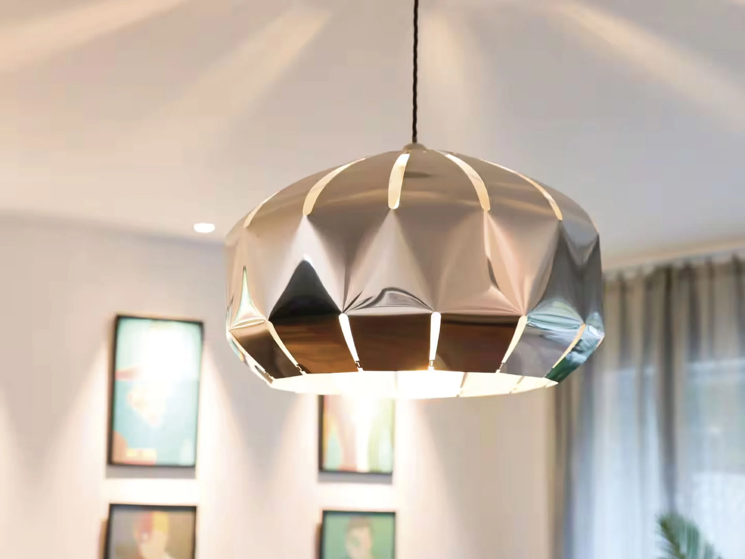 A modern metallic pendant light hanging from the ceiling with a reflection of two large abstract art pieces on the wall behind it.