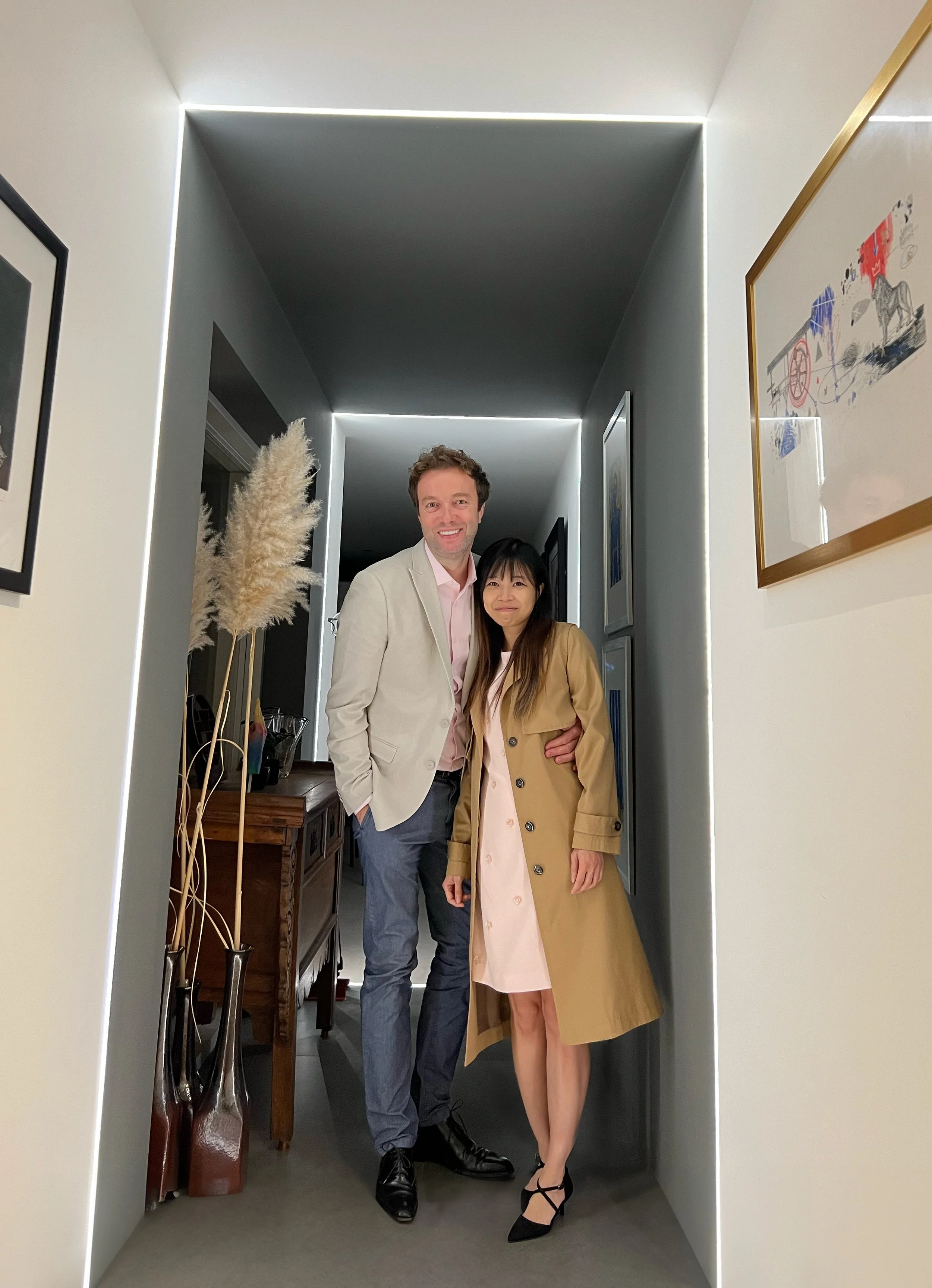 A happy man and woman standing in an entryway, smiling at the camera. The man has curly hair, wearing a beige blazer with a pink shirt and gray pants. The woman has long black hair, wearing a beige trench coat over a pink dress with black high-heeled shoes. The background shows a hallway with framed artwork on the walls and a console table with tall decorative vases.