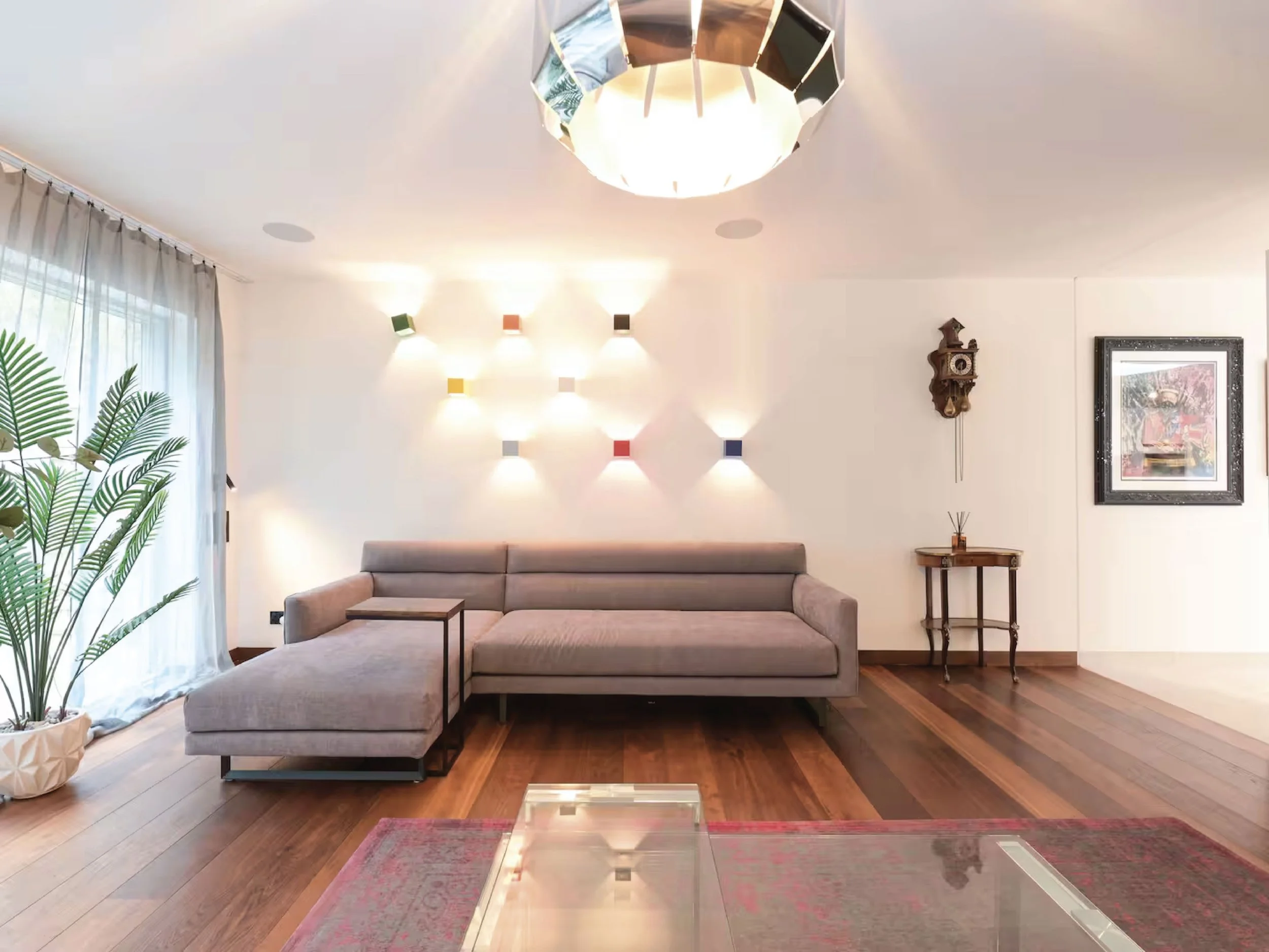 Living room with beige sofa, wooden flooring, large window with white curtains, potted plant, wall art, wall clock, and modern colorful wall-mounted lights.