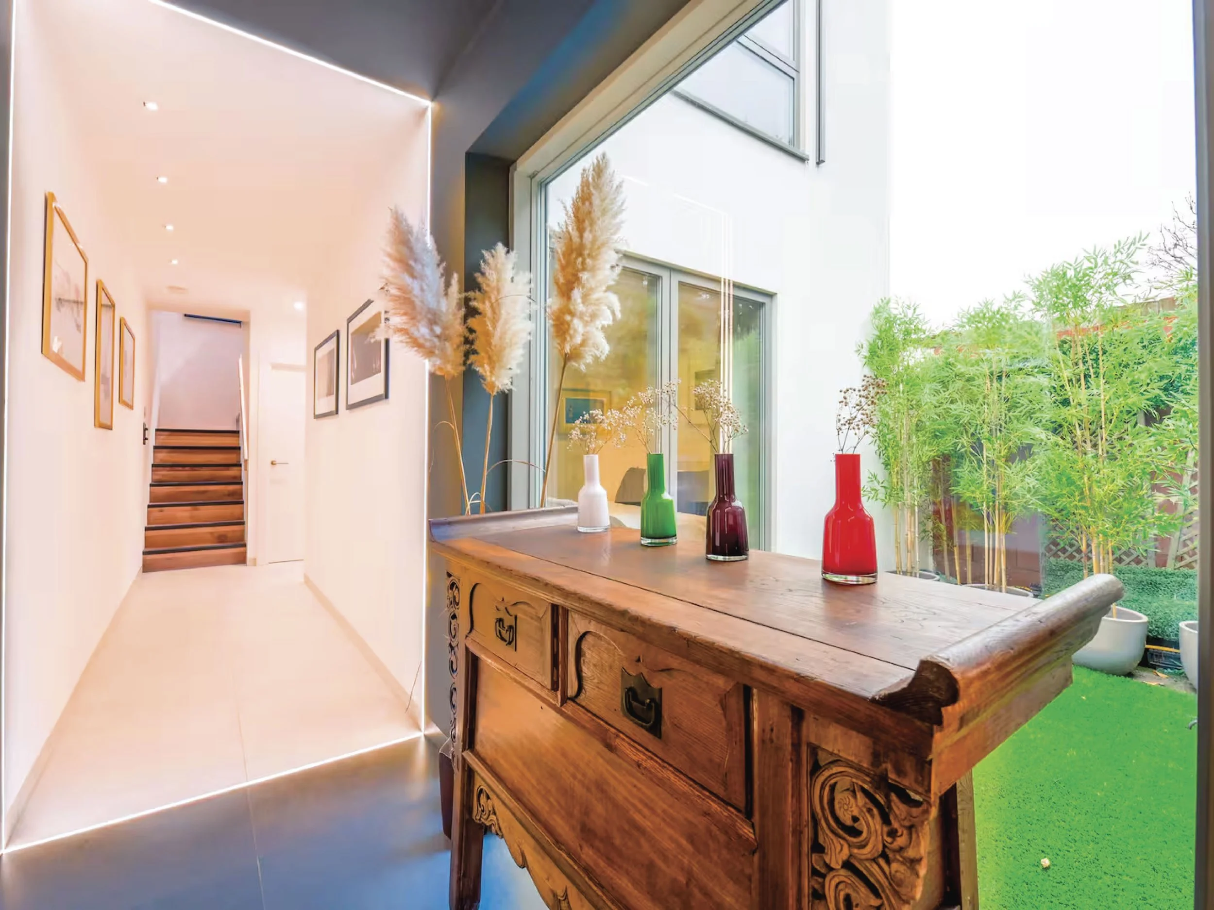 A wooden sideboard with ornate carvings on its edges, decorated with four vases of varying heights and colors holding tall pampas grass and small flowers. A large window behind the sideboard reveals greenery outside, filling the room with natural light.
