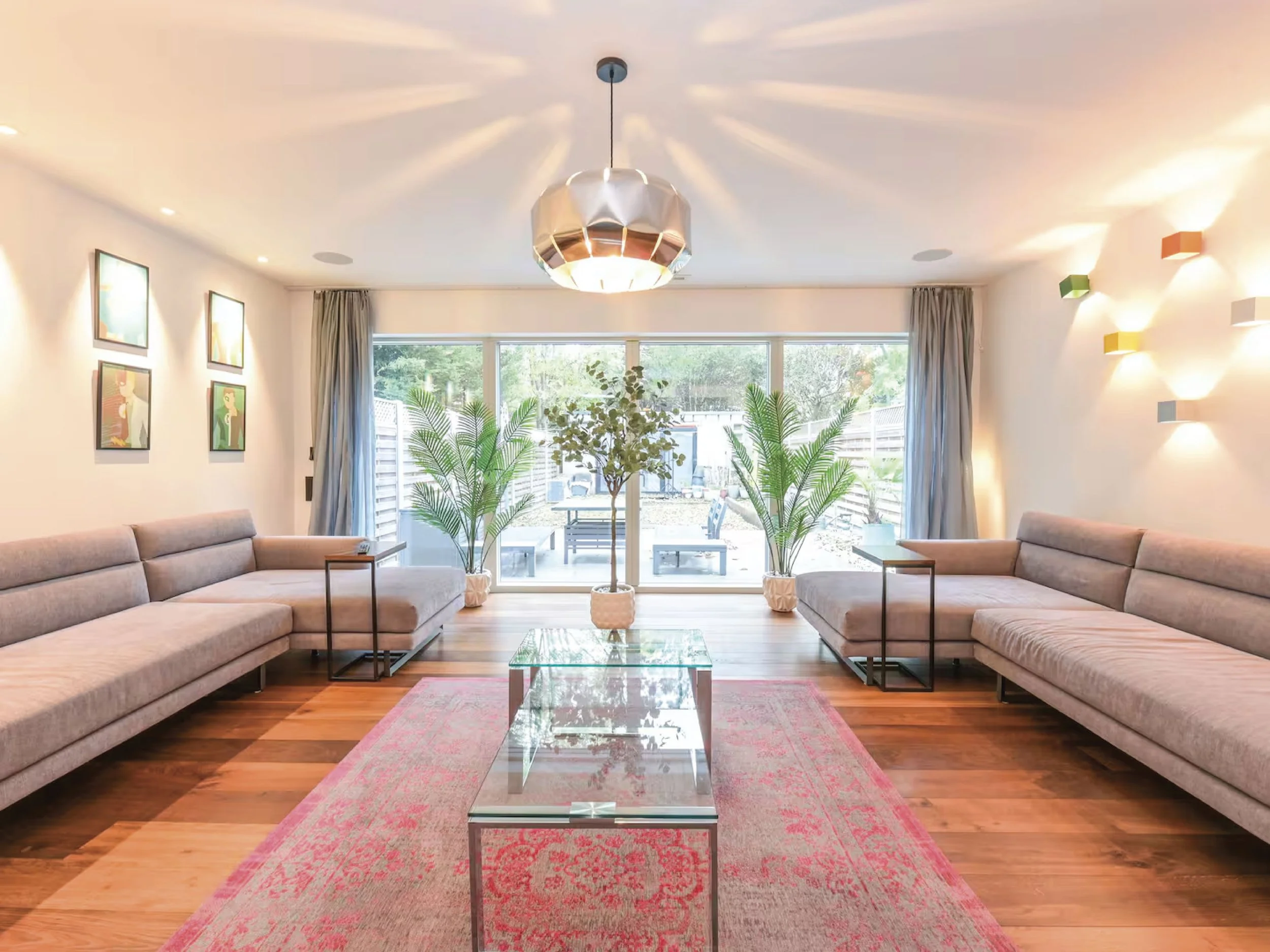 A living room with two large gray sofas, a glass coffee table, and a pink rug. There are three potted plants in the center, artwork on the left wall, and colorful wall-mounted lights on the right wall. A large window with curtains shows a backyard outdoor area.