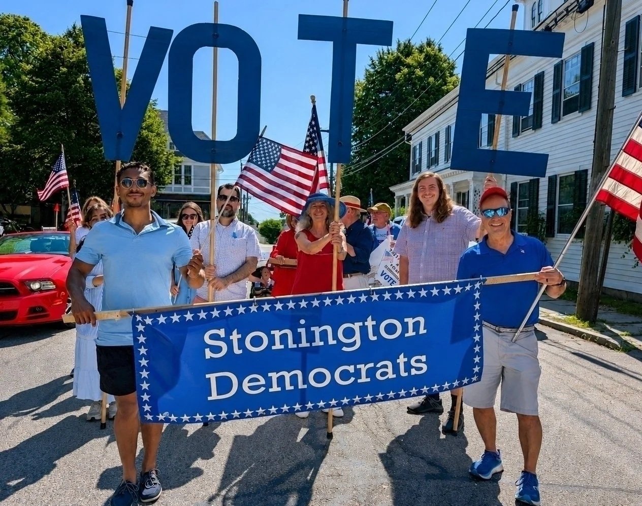 A group of people participating in a parade holding a blue banner that reads "Stonington Democrats," with some also holding American flags and a large "VOTE" sign, walking down a street flanked by houses and cars.