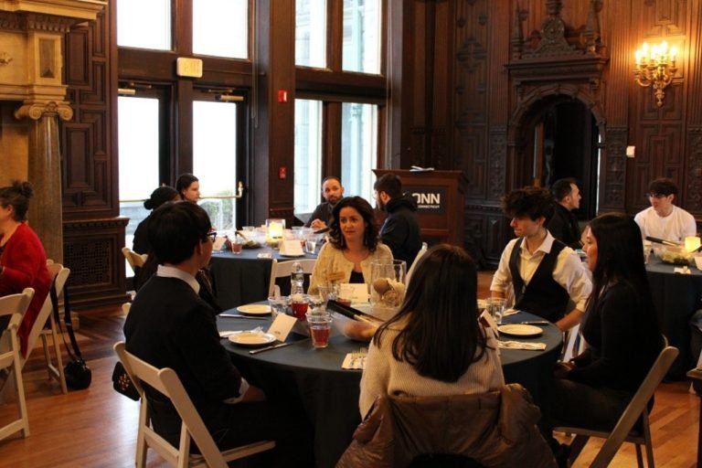 Group of people sitting at round tables in a formal event room with wood-paneled walls and large windows.