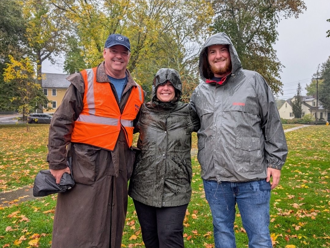 Three people outdoors on a rainy day, wearing rain jackets and smiling.