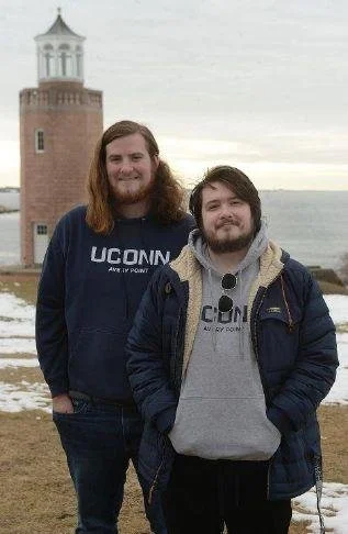 Two young men standing outdoors near water with a lighthouse in the background. Both are smiling and wearing casual clothing, including hoodies and jackets.