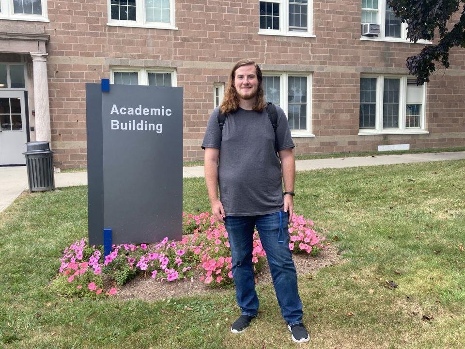 A young man with long hair, wearing a gray t-shirt and blue jeans, standing in front of a sign that says 'Academic Building' on UConn Avery Point. There are pink flowers and a brick building with multiple windows in the background.