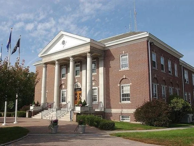 Stonington Town Hall building with white columns and flags outside, surrounded by a landscaped lawn and trees.