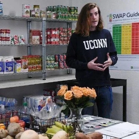 Woman with shoulder-length hair wearing a black UCONN sweatshirt, standing in front of shelves of canned and boxed food items, with a table of vegetables and a vase of orange roses in the foreground.